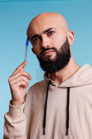 Photo for Puzzled arab man in thoughts holding pen to head while looking away. Young bald bearded person rubbing temple while planning and solving problem with uncertain expression - Royalty Free Image