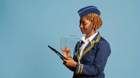 Photo for Female aircrew member using diigtal tablet on camera, scrolling on online website in studio. Young adult with flying uniform looking at gadget screen, browsing social media internet page. - Royalty Free Image