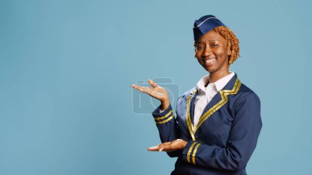 Photo for African american woman advertising something in studio, pointing beside her sideways. Professional stewardess doing ad campaign, point to left or right sides over blue background. - Royalty Free Image