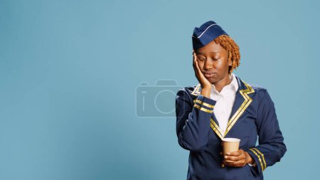 Photo for Air hostess feeling tired drinking coffee and yawning, exhausted aviation worker wearing flying uniform in studio. Stewardess falling asleep and enjoying cup of caffeine beverage. - Royalty Free Image