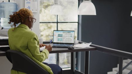 Téléchargez les photos : Africaine américaine analyse les données en ligne sur ordinateur, en regardant le rapport d'analyse à l'écran. Croissance et développement des entreprises dans l'espace de coworking, statistiques économiques. - en image libre de droit
