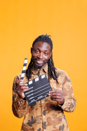 Téléchargez les photos : Portrait d'un homme afro-américain souriant tenant un clapet de film debout en studio sur fond jaune. Jeune adulte donnant audition pour la vidéo de divertissement. Concept cinématographique - en image libre de droit