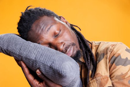 Foto de Adulto joven agotado que pone la cabeza en una almohada cómoda para dormir y tomar la siesta. hombre afroamericano tratando de descansar después de pasar la hora de acostarse, durmiendo en el estudio sobre fondo amarillo - Imagen libre de derechos