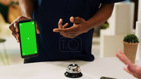 Photo for Male tourist holding phone with greenscreen, pointing at mockup chromakey template on display. African american guest holding screen with isolated copyspace at reception front desk. - Royalty Free Image