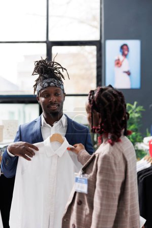 Foto de Showroom manager ayudando a los clientes con estilo con camisa blanca en la boutique moderna. Cliente afroamericano que compra ropa y accesorios de moda para aumentar el guardarropa. Concepto de moda - Imagen libre de derechos