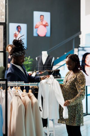 Photo for Boutique employee helping customer with casual wear, looking at white shirt discussing fabric in clothing store. African american client buying maternity clothes and trendy merchandise - Royalty Free Image