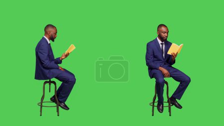 Téléchargez les photos : Homme afro-américain lisant un livre de roman assis sur une chaise, appréciant le conte comme littérature en studio. Jeune en costume ayant des pages de conférences sur écran vert, activité de loisirs à la caméra. - en image libre de droit