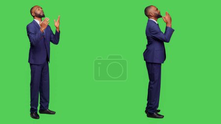 Photo for Spiritual office worker praying to god for luck on camera, wearing business suit over full body greenscreen. Corporate employee holding hands in a prayer, religious man with beliefs. - Royalty Free Image
