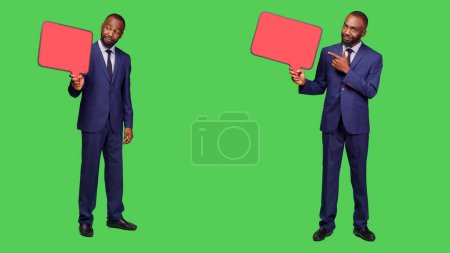 Photo for Young entrepreneur showing empty speech bubble, using blank isolated copyapce for advertisement on camera. Office worker in suit using red icon billboard for ad on green screen backdrop. - Royalty Free Image