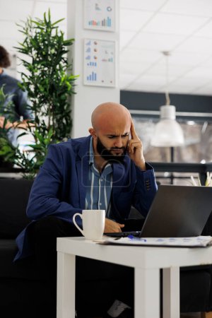 Téléchargez les photos : Entreprise fatiguée arabe employé souffrant de migraine tout en travaillant sur ordinateur portable d'entreprise au bureau. Démarrage épuisé directeur exécutif résoudre la tâche de l'entreprise dans l'espace de coworking - en image libre de droit