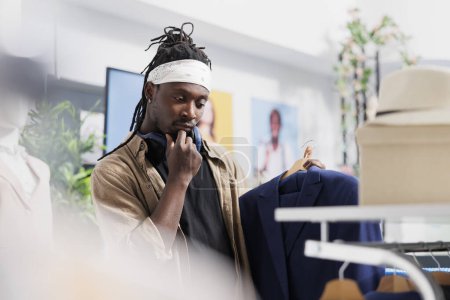 Buyer shopping for jacket and thinking about choice in clothing store. African american man holding blazer on hanger while rubbing chin and looking at apparel style in mall
