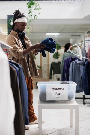 Foto de Cliente de la tienda de ropa que da prendas al banco de donaciones mientras compra ropa. Hombre afroamericano poniendo ropa en contenedor de plástico de caridad para los pobres en boutique de moda - Imagen libre de derechos
