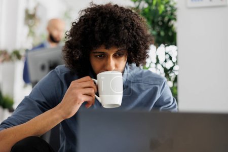 Foto de Empleado árabe gestiona SEO optimización en el ordenador portátil mientras bebe café en la oficina corporativa. Joven profesional sosteniendo taza de té y analizando datos analíticos de informes en el espacio de coworking - Imagen libre de derechos