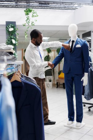 Photo for Boutique african american employee checking mannequin wearing stylish jacket and trousers. Man adjusting formal male trendy suit on dummy model in department shopping mall - Royalty Free Image