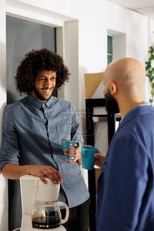 Téléchargez les photos : Deux collègues arabes rient et discutent occasionnellement tout en buvant du café dans le bureau de l'entreprise. Démarrer des employés de l'entreprise bavarder pendant la pause dans l'espace de coworking - en image libre de droit