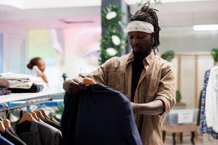 Foto de Shopper sostiene la chaqueta mientras examina la tela y el tamaño antes de comprar ropa en la tienda de ropa. Hombre afroamericano revisando chaqueta casual en percha en boutique centro comercial - Imagen libre de derechos