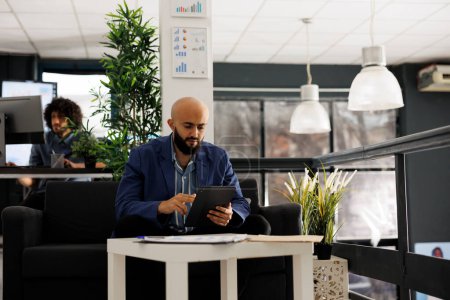 Téléchargez les photos : Homme d'affaires arabe utilisant tablette pour le travail numérique dans le bureau d'affaires. Employé de l'entreprise défilant dans les médias sociaux pendant la pause tout en étant assis sur le canapé dans l'espace de coworking - en image libre de droit