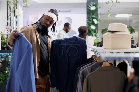 Photo for Buyer deciding between two jackets while choosing formal wear in clothing store. African american man holding blazers on hangers while examining style and fabric in shopping center - Royalty Free Image