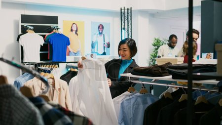 Photo for Asian customer examining formal clothes on hangers and buying new collections from retail shop. Young adult visiting clothing store to try on trendy modern merchandise, commercial activity. - Royalty Free Image