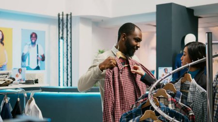 Photo for African american man checking material of clothes and consulting store assistant before buying fashionable merchandise. Male client talking to asian woman in mall department store. - Royalty Free Image