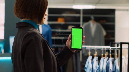 Photo for Young assistant using blank greenscreen template, showing chroma key display in trendy clothing store. Asian worker using isolated mockup copyspace on modern smartphone, shopping center. - Royalty Free Image