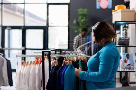 Foto de Cliente afroamericano mirando perchas llenas de ropa de moda en la boutique moderna, comprando mercancía de moda. Mujer Shopaholic comprobando nueva colección de moda, analizando la tela - Imagen libre de derechos