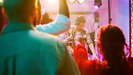Photo for Persons dancing with DJ at nightclub, young adult mixing funky music for group of friends on dance floor. Happy people partying and having fun at celebration with disco lights. Handheld shot. - Royalty Free Image