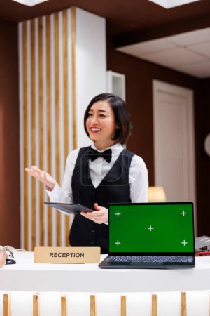 Photo for Hotel worker with green screen on desk, welcoming people and working on registration with tablet. Asian receptionist having laptop with chroma key display and isolated mockup screen. - Royalty Free Image