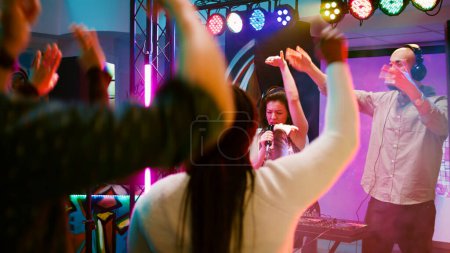 Téléchargez les photos : Femme heureuse faisant du karaoké à la soirée dansante, chantant des chansons au microphone sur scène avec DJ mixant de la musique. Des gens joyeux dansant en s'amusant au rassemblement social, au divertissement. Coup de main. - en image libre de droit