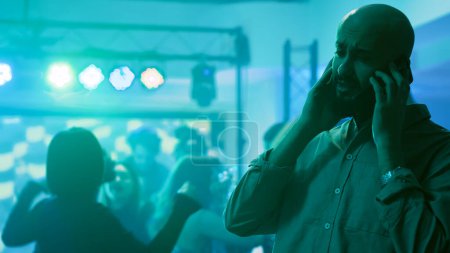 Photo for Male adult chatting on phone at party, trying to talk to someone on mobile phone remote call in discotheque. Young person attending dance party at club, answering smartphone. Tripod shot. - Royalty Free Image