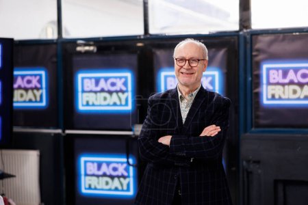 Photo for Happy senior man customer standing with crossed arms against neon Black Friday banners on wall in retail store, smiling at camera. People enjoying shopping during big sales - Royalty Free Image