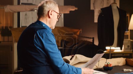 Photo for Old craftsman making clothing items, looking at drawings in tailor workshop to create custom made clothes. Experienced couturier working with tools and skill, craftsmanship. Handheld shot. - Royalty Free Image