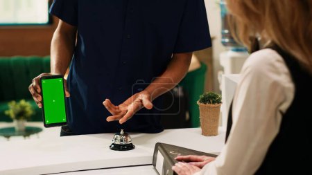 Photo for Hotel guest showing greenscreen on phone, pointing at isolated display on smartphone. African american man holding device with blank chromakey template at reception front desk. - Royalty Free Image