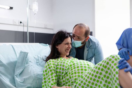 Foto de Mujer embarazada empujando y gritando durante el parto, recibiendo apoyo médico del marido y del equipo del hospital. La futura madre tiene contracciones dolorosas durante el parto del bebé en la clínica de maternidad - Imagen libre de derechos