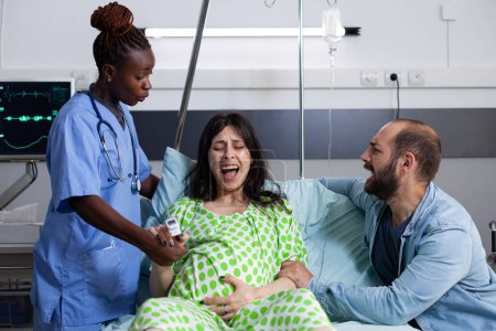 Foto de Mujer con dolor de embarazo recibiendo asistencia médica de enfermera afroamericana en la sala de hospital. Futuro padre de pie junto a la esposa embarazada apoyándola y consolándola sobre el parto - Imagen libre de derechos