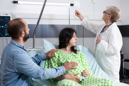 Foto de Médico mayor revisando bolsa de goteo intravenoso, explicando la cirugía de cesárea a la mujer embarazada en la sala de hospital. Mujer con embarazo acostada en la cama discutiendo la maternidad con el futuro padre - Imagen libre de derechos