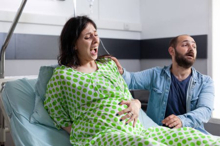 Foto de Marido aterrorizado gritando por el médico mientras la mujer embarazada tiene contracciones dolorosas en la sala de hospital. Paciente con embarazo que entra en trabajo de parto, pidiendo asistencia médica antes de la cirugía - Imagen libre de derechos