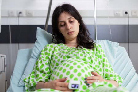 Téléchargez les photos : Femme enceinte tenant la main sur le ventre, couchée au lit se préparant pour l'accouchement dans la salle d'hôpital. Patiente caucasienne enceinte portant un oxymètre, enceinte en maternité - en image libre de droit