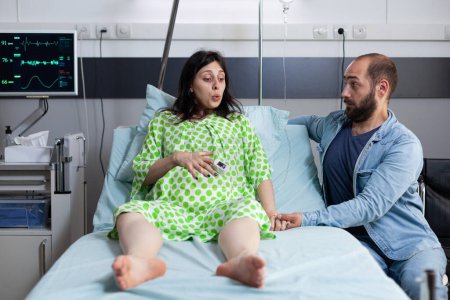 Foto de Paciente embarazada sentada en la cama de la sala de hospital con el marido a su lado esperando el parto. Mujer que se prepara para el trabajo de parto que tiene contracciones del dolor, pidiendo ginecólogo para venir y comenzar la cirugía - Imagen libre de derechos