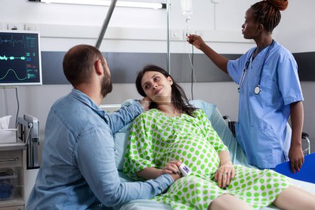 Foto de Pareja con embarazo esperando el parto en la sala de hospital. Enfermera afroamericana revisando bolsa de goteo IV mientras mujer embarazada acostada en la cama y marido reconfortante sobre la maternidad - Imagen libre de derechos