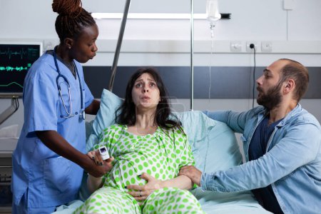 Foto de Mujer embarazada en trabajo de parto que sufre de contracciones dolorosas que yacen en la cama de la sala de hospital. Enfermera y esposo afroamericanos apoyando el embarazo paciente, tomados de la mano para asistencia médica - Imagen libre de derechos