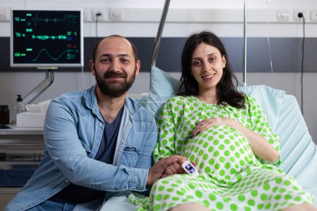 Foto de Alegre futuros padres que se preparan para el parto del bebé en la sala del hospital, esperando a que el médico venga y comience la cirugía de cesárea. Mujer embarazada acostada en la cama en la clínica de maternidad. Concepto de parto - Imagen libre de derechos