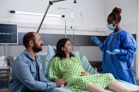 Afrikanisch-amerikanischer Chirurg mit Schutzhandschuhen und Maske diskutiert mit schwangerer Frau die Entbindungschirurgie auf der Krankenhausstation. Frau mit Schwangerschaft liegt mit Wehen im Bett