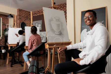Foto de Retrato de un joven afroamericano feliz estudiante de la escuela de arte sentado en el caballete sonriendo a la cámara, aprendiendo dibujo a lápiz en clase. Positivo negro chico disfrutando creativo hobby con amigos - Imagen libre de derechos