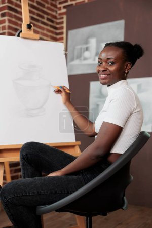 Foto de Feliz positivo afroamericano millennial estudiante obtener placer en el dibujo mientras estudia en la escuela de arte. Mujer joven sonriente sentada en el caballete dibujando con lápiz sobre lienzo durante el taller - Imagen libre de derechos