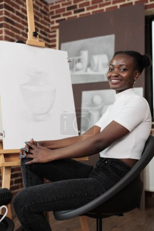 Foto de Retrato de niña afroamericana feliz sonriente sentada en el caballete sintiéndose satisfecha con la obra de arte que creó durante la clase de dibujo. Joven mujer alegre aprendiendo cosas nuevas y divirtiéndose - Imagen libre de derechos