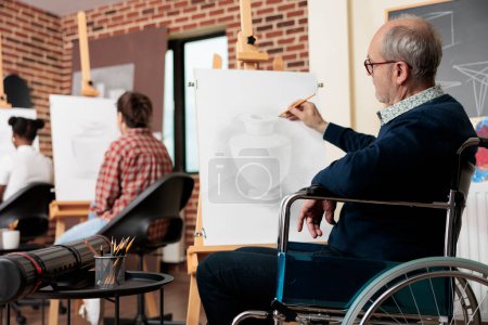 Foto de Arte terapia para discapacidades físicas. Hombre mayor usuario de silla de ruedas dibujo sobre lienzo, persona con discapacidad que asiste a la clase de grupo terapéutico creativo. Actividades para personas con movilidad reducida - Imagen libre de derechos