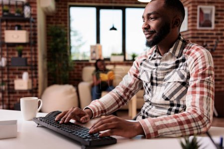 Téléchargez les photos : Souriant afro-américain pigiste travaillant à distance dactylographier et regarder l'écran d'ordinateur pendant colocataire se détendre sur le canapé. Programmeur masculin utilisant PC pour développer des logiciels à la maison lecture e-mails. - en image libre de droit