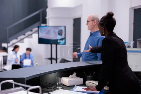 Photo for Reception worker telling sick old man to take a seat in waiting area during checkup visit appointment in hospital. Elderly man having medical consultation with physician doctor, health care service - Royalty Free Image