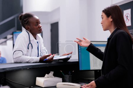 Photo for Asian receptionist talking with specialist doctor at hospital reception counter discussing patient symptoms after consultation. Medical staff planning medication treatment to cure disease - Royalty Free Image